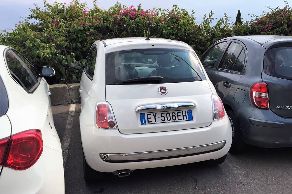 Een kleine witte Fiat 500 staat geparkeerd in een piepklein gaatje. Autorijden op Sicilië gaat bepaald niet vanzelf.