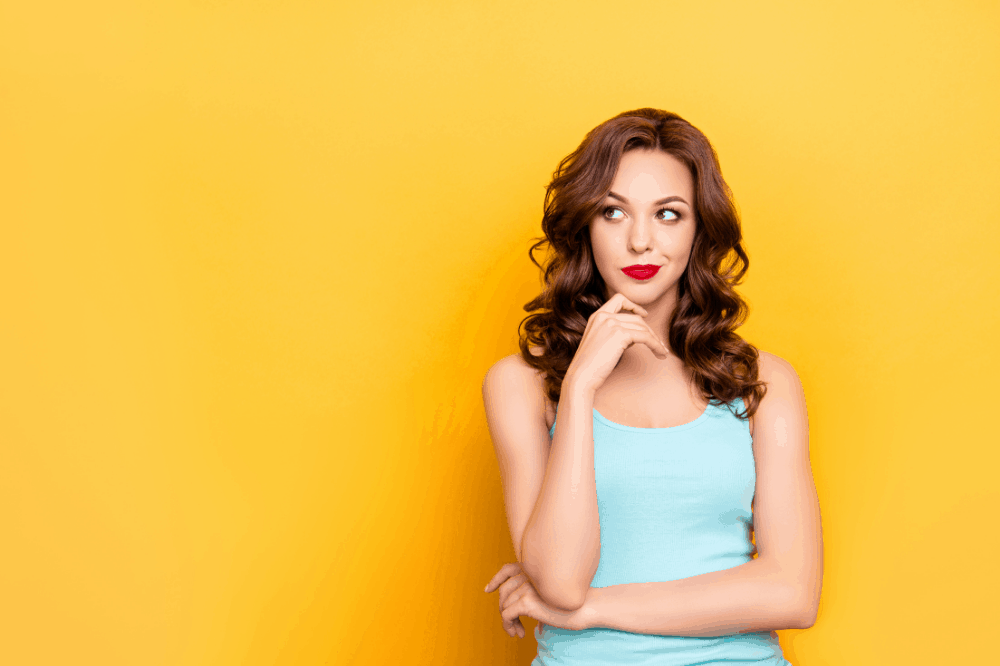 Een witte vrouw met bruine krullen kijkt twijfelachtig opzij, ze staat voor een gele muur, wellicht gaat haar twijfel over deelname aan Zin in Aflossen, de aflostraining van Fuck Die Studieschuld