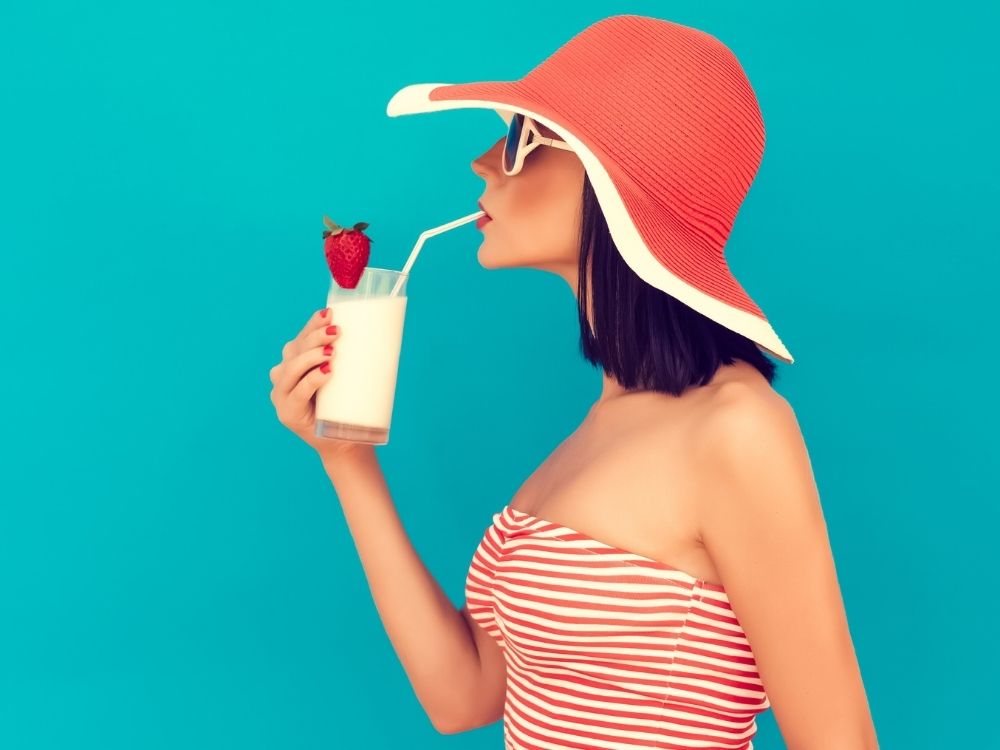 Zijaanzicht van een witte vrouw met zonnebril en hoed, ze drinkt een milkshake (ze is klaar voor de zomer en wellicht ook voor de Zomerserie van Fuck Die Studieschuld)