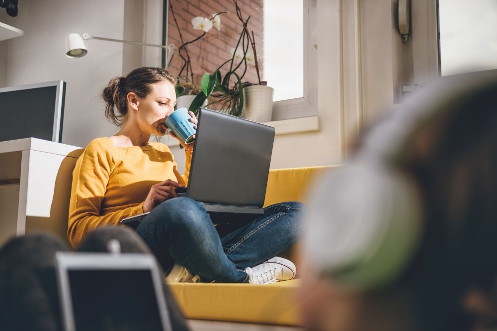 Een jonge vrouw in een gele trui zit op een gele bank met op haar schoot een laptop