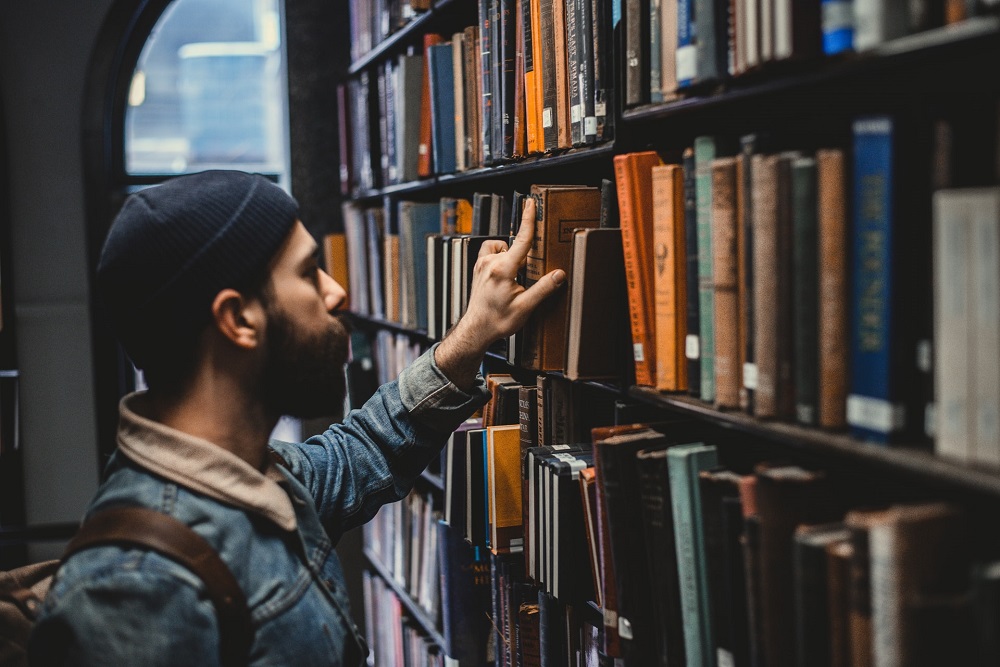 Een man met spijkerjas en muts zoekt in een bibliotheek naar een boek (wellicht zoekt hij naar het geheim van het sneller aflossen van je studieschuld)