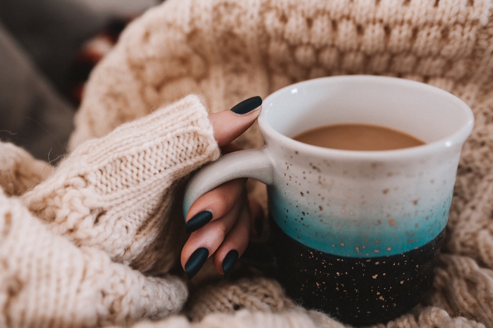 Een vrouw in een warme trui met een kop warme koffie (een goede strategie voor als je je energierekening laag wilt houden)