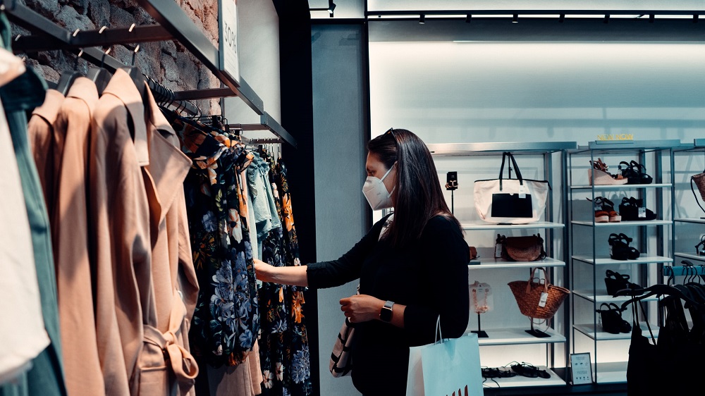 Een vrouw met zwarte haren en een zwarte jas bekijkt kleding in een kledingwinkel (pas op met dingen kopen waarvan je denkt dat je ze echt nodig hebt terwijl je er geen geld voor hebt)