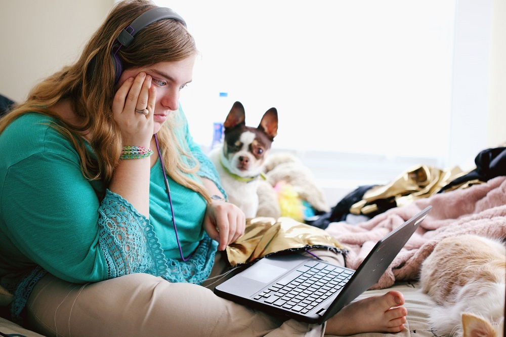 Een blonde vrouw zit met een hondje op een bed, ze staart zorgelijk naar het scherm van haar laptop (wellicht leest ze dat de rente van DUO stijgt)