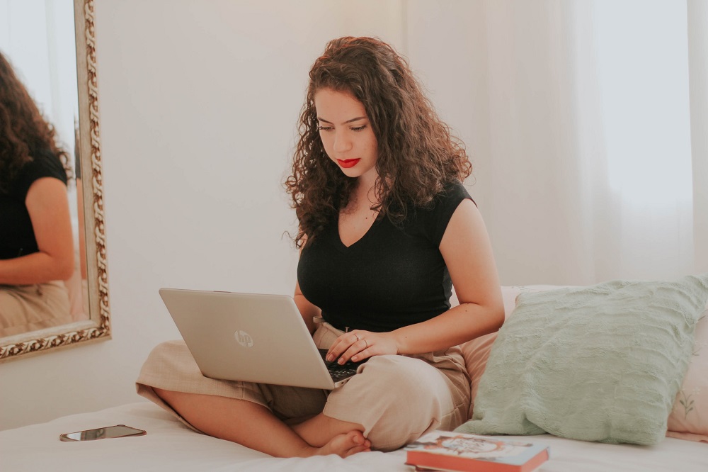 Een vrouw met donkere krullen werkt al zittend op een bed op haar laptop (wellicht heeft ze zin om te bloggen)