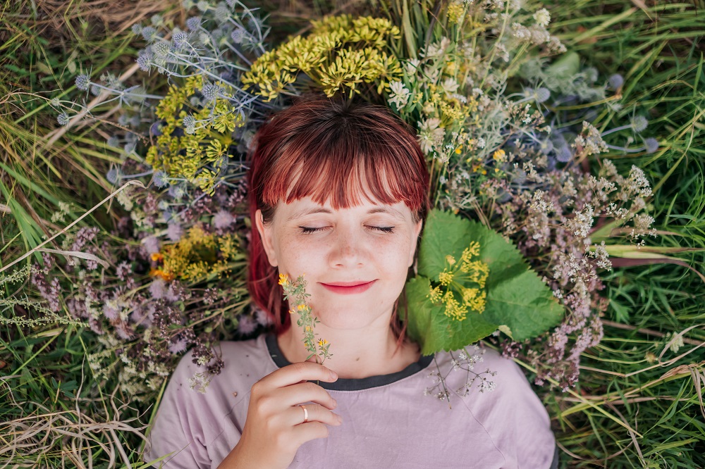 Een jonge vrouw ligt op haar rug in het gras tussen de bloemen, haar ogen zijn dicht, ze oogt kalm en gelukzalig (doe eens wat vaker wat je zelf wilt)