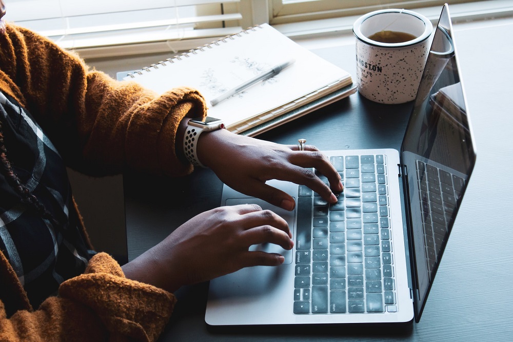 Een zwarte vrouw werkt op een laptop, naast haar een notitieblok en een kop thee (ze is wellicht aan het budgetteren)