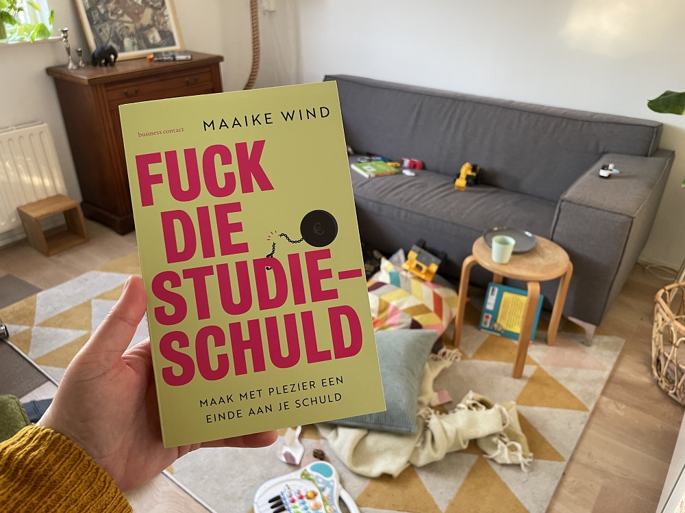 Een foto van mijn boek Fuck die studieschuld met op de achtergrond een rommelige woonkamer (ik verbaas me over dat schrijven zo wonderlijk is)