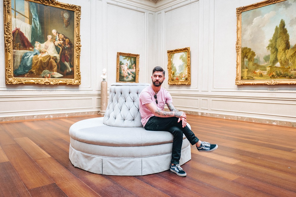 Een witte man met tatoeages zit op een bankje in een museum, om hem heen hangen schilderijen (wellicht had hij zin om zichzelf te trakteren)