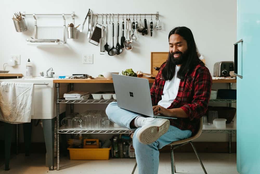 Een man met lang haar zit in een keuken op een stoel en kijkt glimlachend naar de laptop op zijn schoot (hij heeft wellicht voordelen van het aflossen op de korte termijn ontdekt)